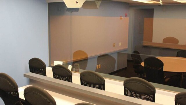 Viewing room at focus group facility showing 2 tiers of desktop with multiple office chairs, blue walls, wall mounted speakers and the one-way glass.